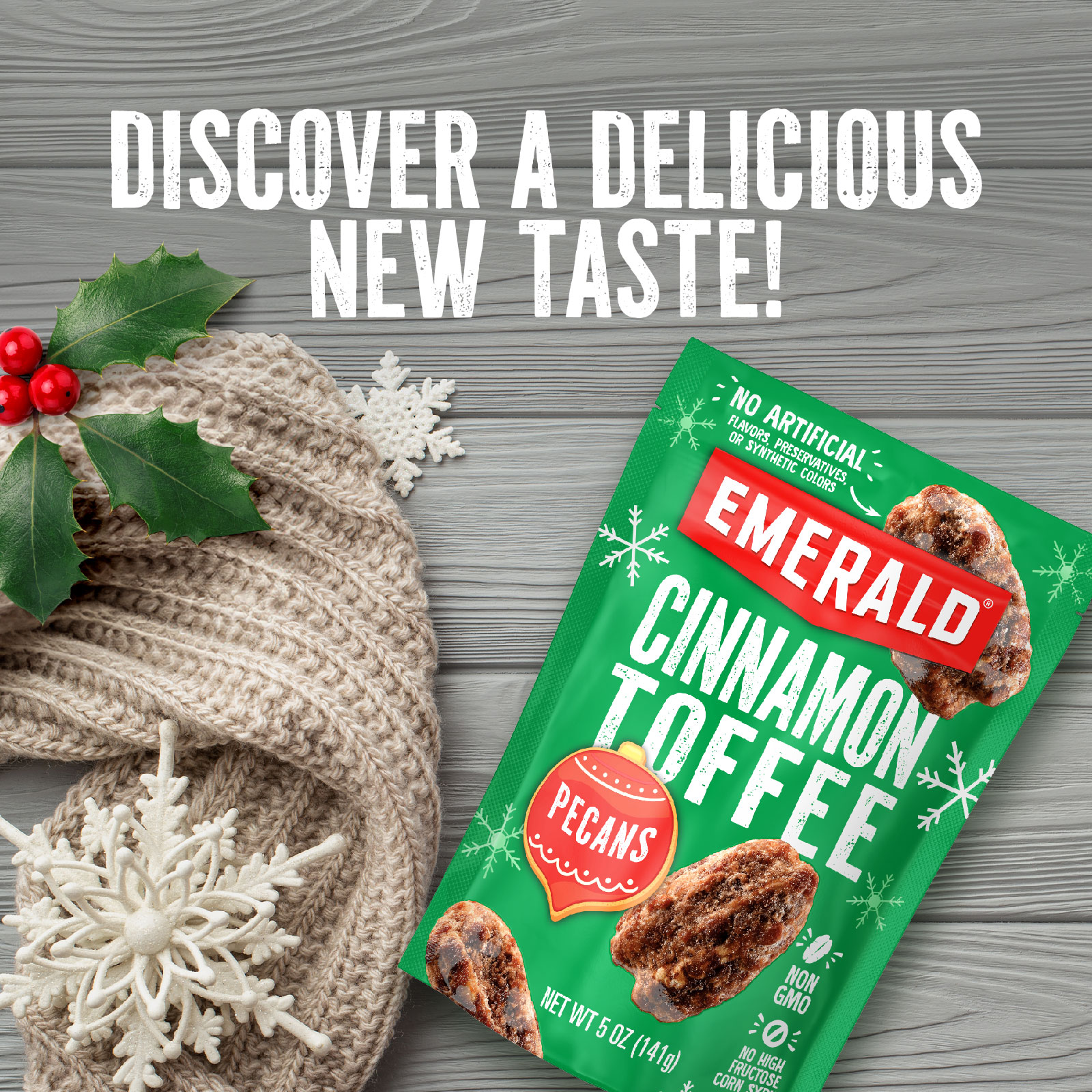 A packet of Emerald Cinnamon Toffee Pecans is displayed on a wooden surface next to a knitted scarf, red berries, green leaves, and a white snowflake ornament. Text above reads, Discover a delicious new taste!.