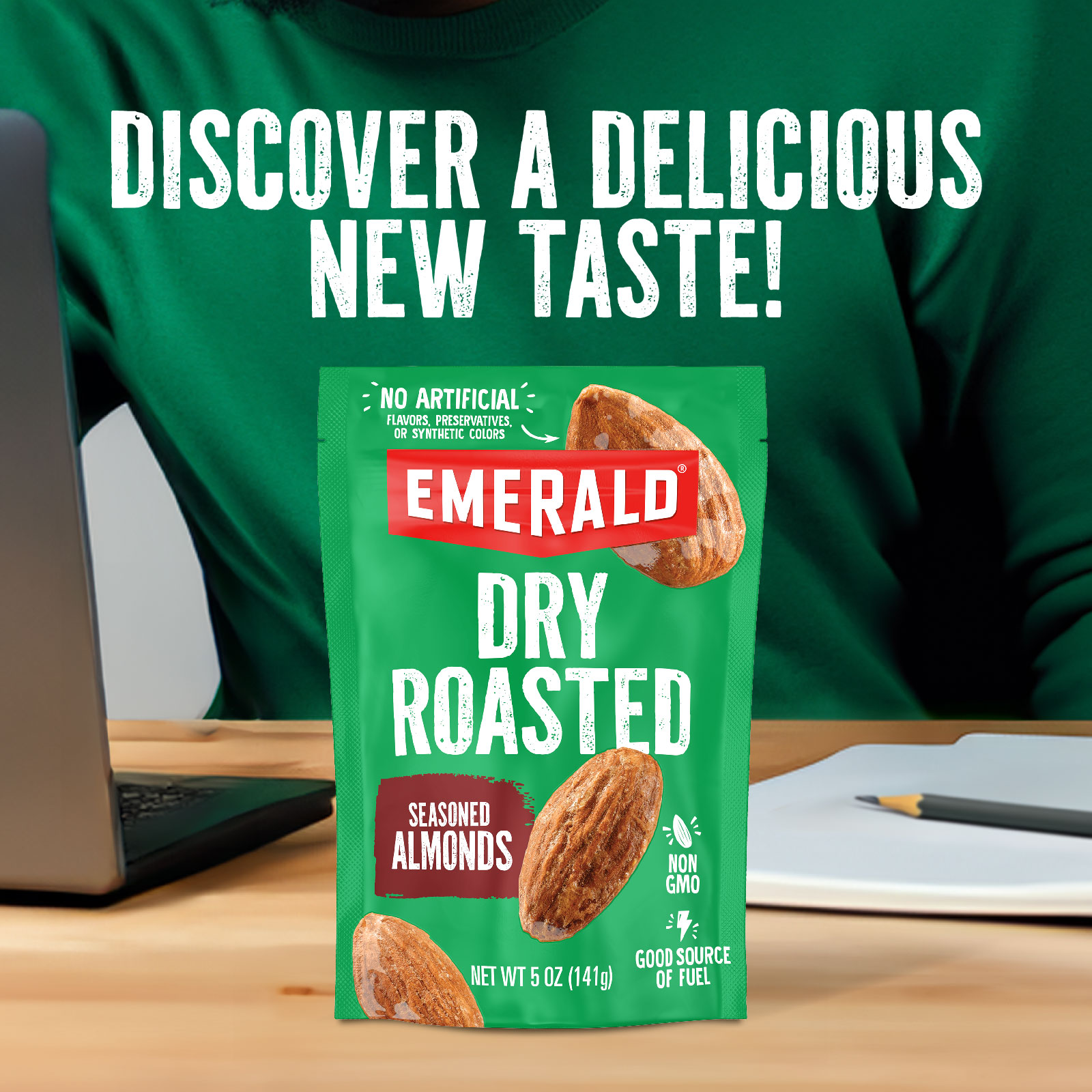 A bright green package of Emerald Dry Roasted Seasoned Almonds is placed on a desk beside an open laptop. A person in a green sweater is in the background. Text above reads, Discover a delicious new taste!.